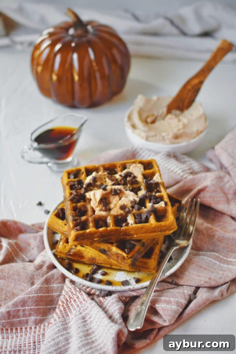 A freshly cooked pumpkin waffle, golden and crisp, resting on a plate, topped with a dollop of cinnamon butter, a generous pour of maple syrup, and a scattering of chocolate chips.
