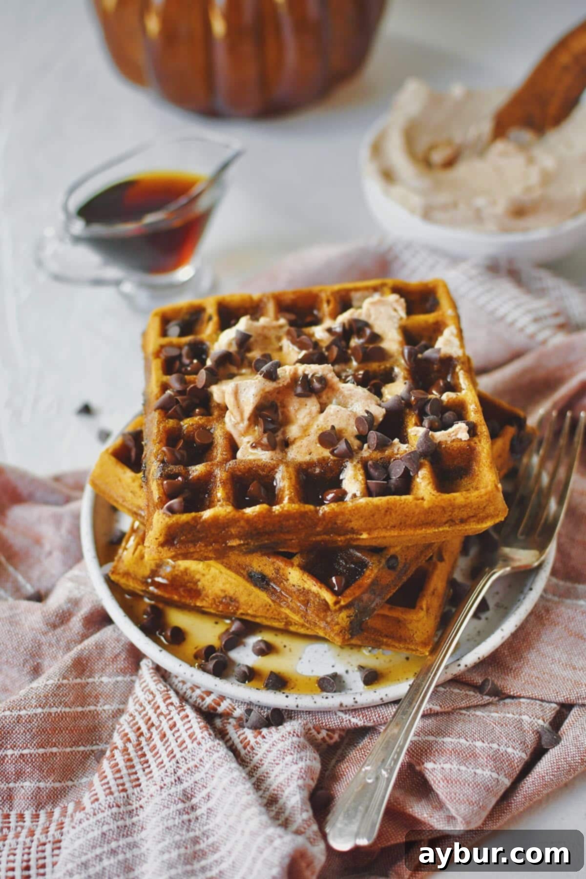 A close-up of a stack of fluffy Pumpkin Waffles on a white plate, showcasing the melted chocolate chips, cinnamon butter, and rich maple syrup cascading down the sides.