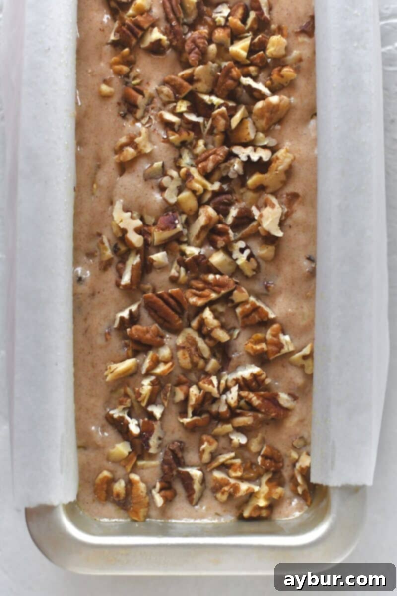 Banana bread batter in a prepared loaf pan with parchment liner, before baking, topped with nuts for a crunchy crust.