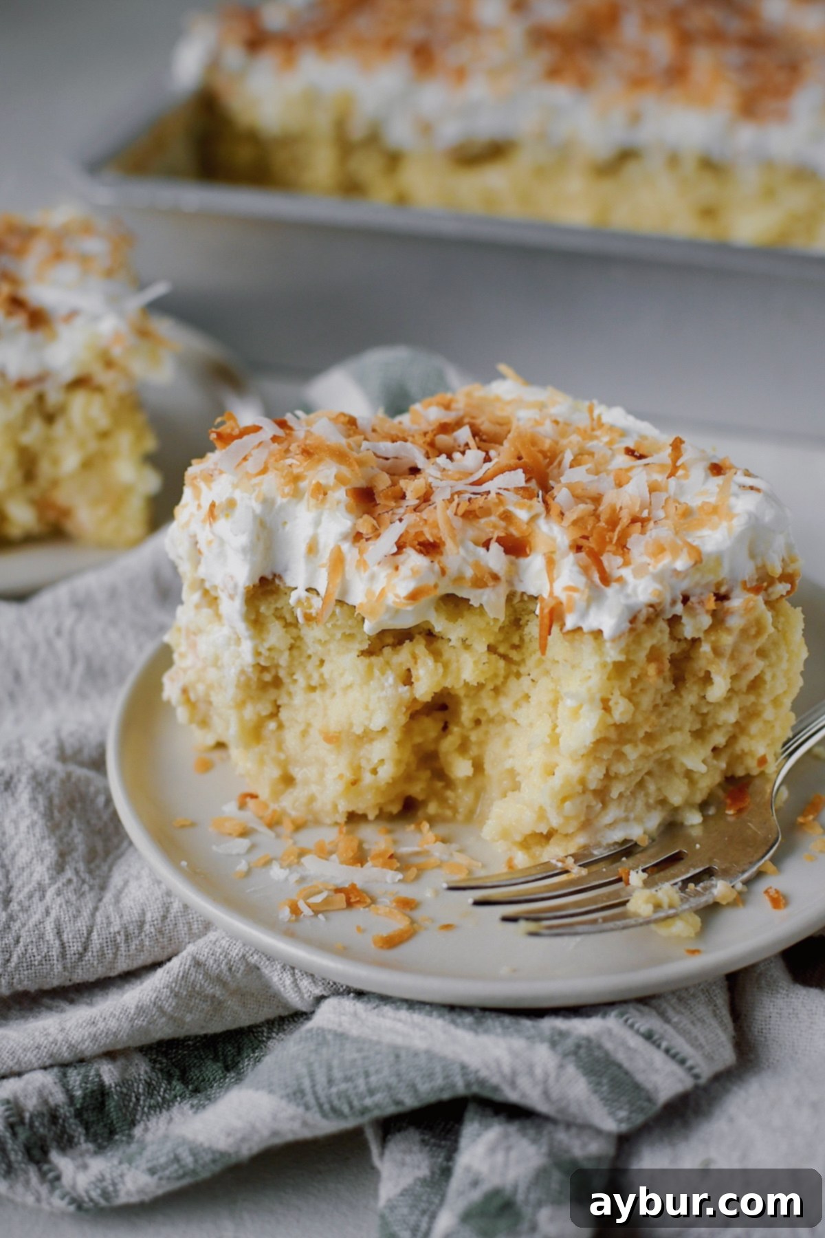 A slice of Coconut Tres Leches on a plate, with a few bites having been taken from it. Fork on the side of the plate.