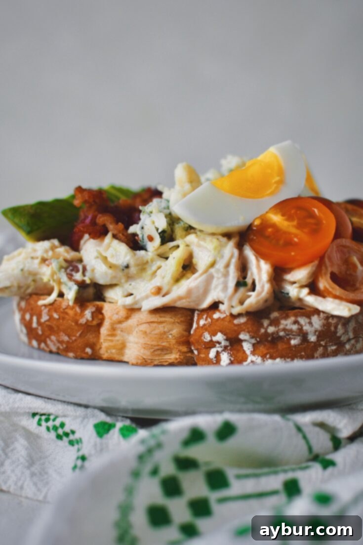 Side view of dressed Chicken Salad on toasted white bread, topped with tomatoes, eggs, blue cheese, bacon, an avocado all lined up like a cobb salad would be.