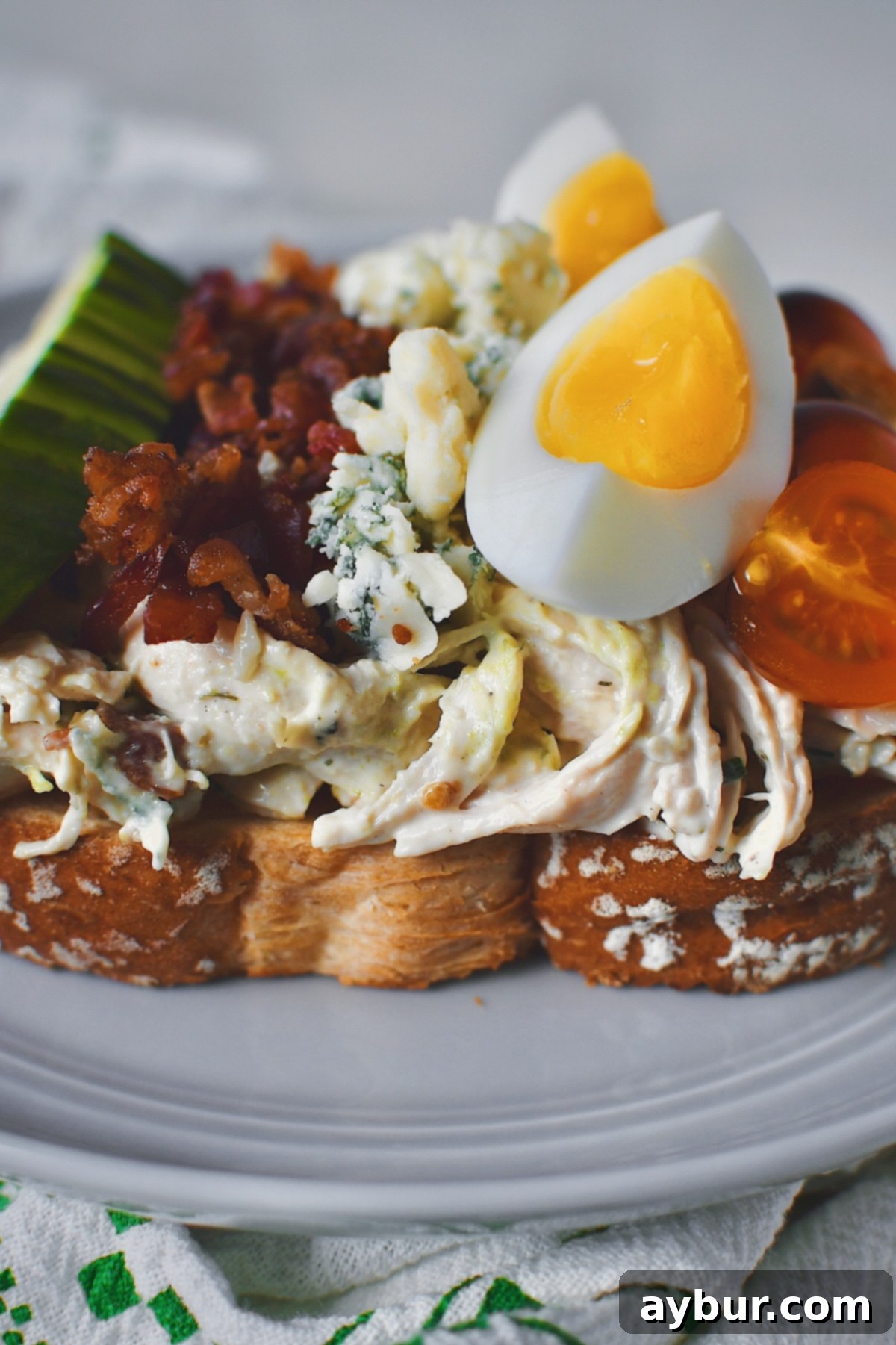 Looking straight on at a dressed Chicken Salad on toasted white bread, topped with tomatoes, eggs, blue cheese, bacon, an avocado all lined up like a cobb salad would be, showcasing the beautiful layers.
