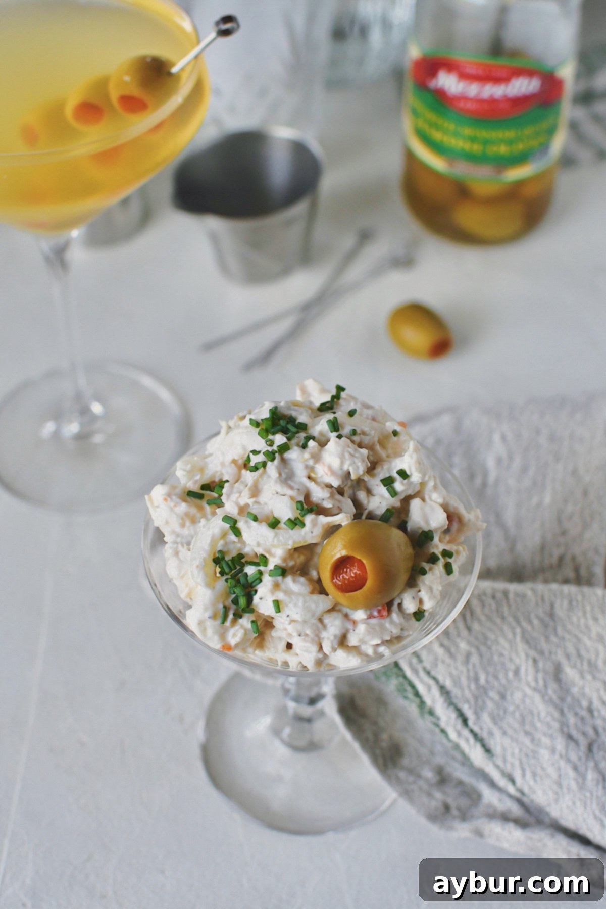 A close-up view of the Martini Chicken Salad, beautifully presented in a martini glass and garnished with an olive and chives, with a chilled martini cocktail blurred in the background.