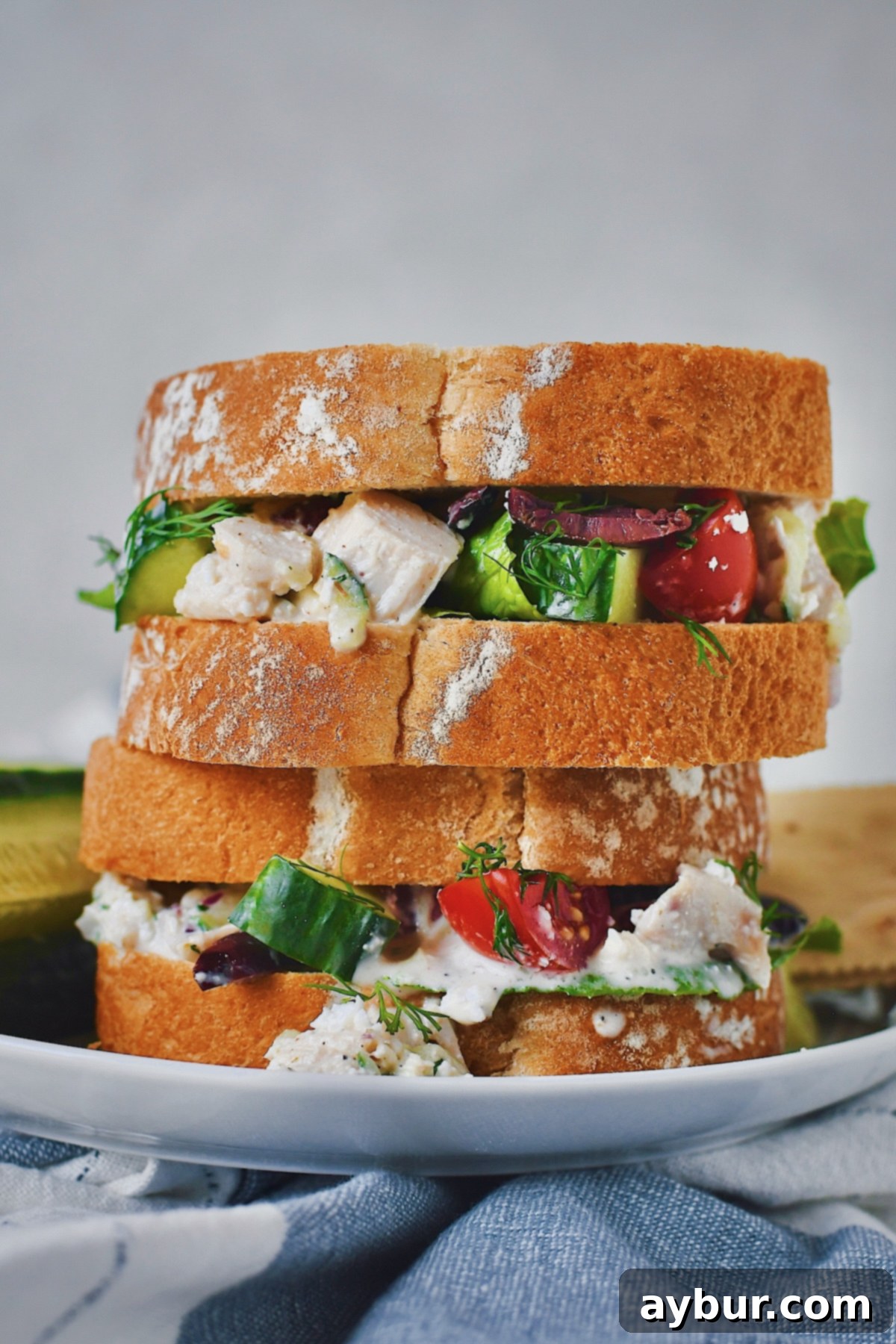 Greek Style Chicken Salad Sandwich, beautifully cut in half and stacked, presented on a plate with refreshing cucumber sticks and crispy crackers.