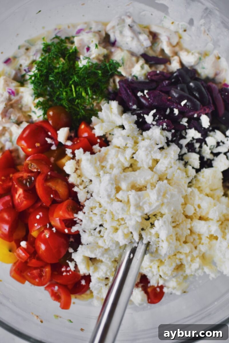 Finishing the chicken salad mixture by folding in vibrant tomatoes, olives, crumbled feta cheese, and fresh dill.
