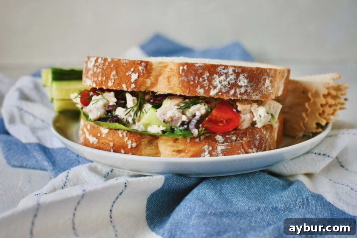 A delicious Greek Style Chicken Salad sandwich served on white bread, presented on a plate with crunchy crackers and refreshing cucumber sticks.