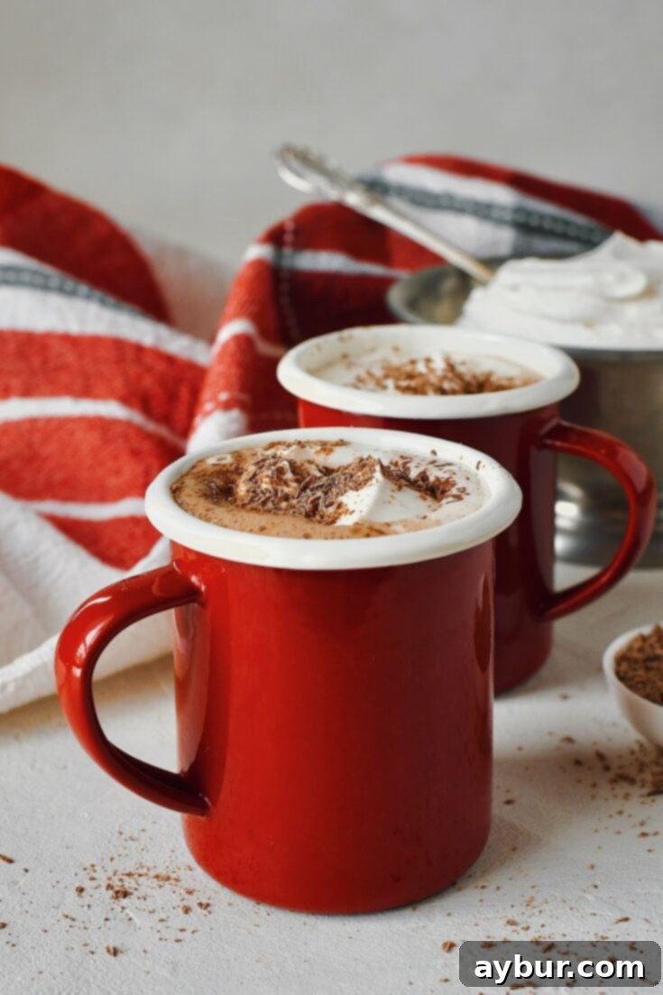 Two mugs of Nutella Hot Chocolate with chocolate shavings on the side, and a bowl of fresh whipped cream behind them, ready for Pinterest sharing.