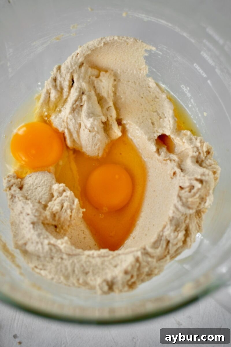 Two large eggs being added to the light and fluffy creamed butter and sugar mixture in a stand mixer, essential for creating a rich cookie dough base.