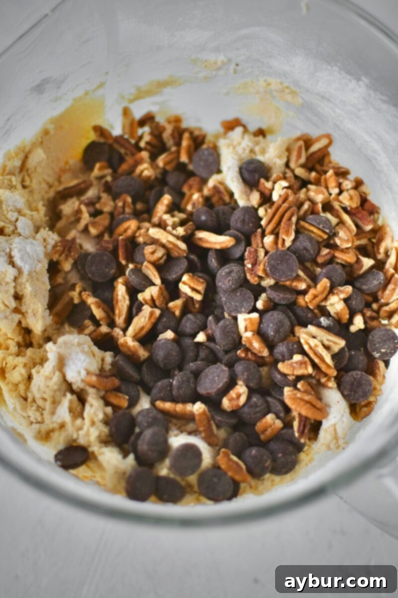 Semisweet chocolate chips and finely chopped pecans being incorporated into the cookie dough by hand, ensuring even distribution and preventing breakage.
