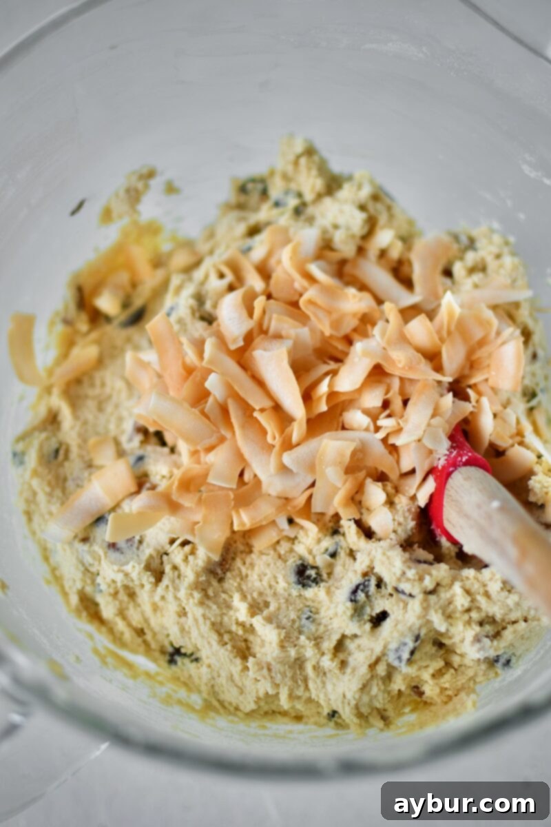 The final step of gently folding in the delicate toasted coconut chips into the cookie dough mixture by hand, preserving their crisp texture.