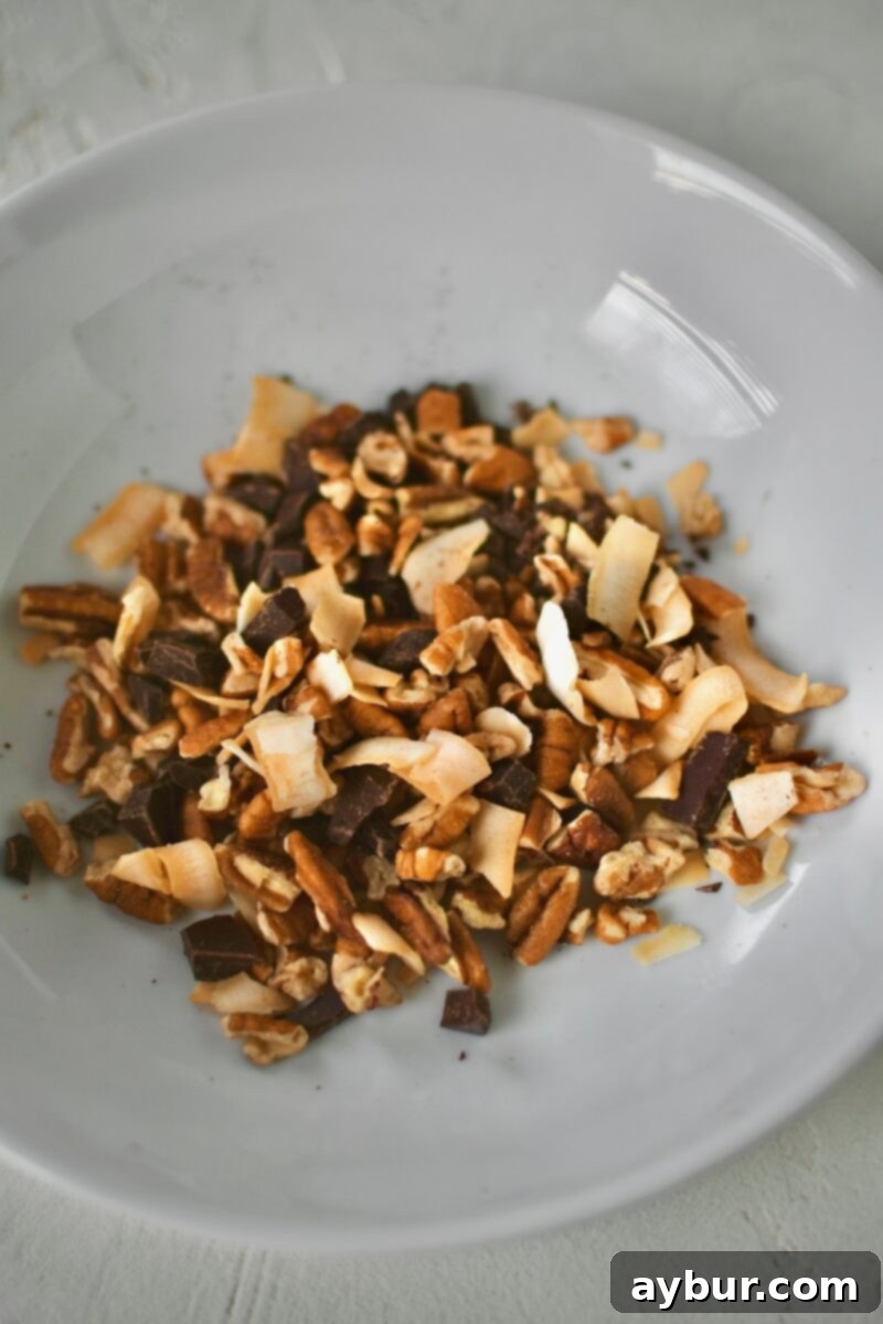 A close-up shot of a bowl brimming with an enticing mixture of extra semisweet chocolate chips, crunchy toasted coconut chips, and chopped pecan pieces, ready to be used as a cookie topping.