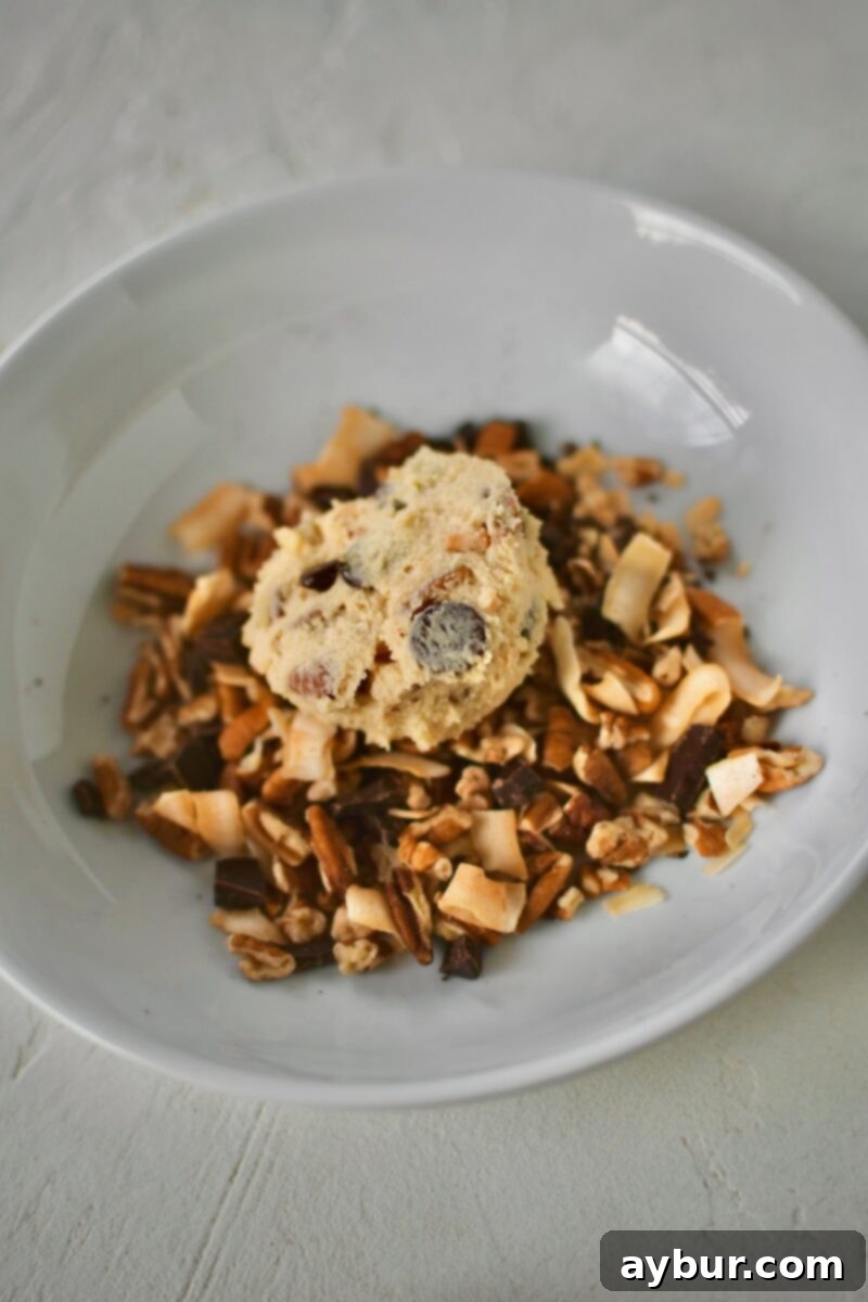 The artistic process of carefully dipping a perfectly scooped cookie dough ball into the rich topping mixture, ensuring a generous coating of chocolate, coconut, and pecans.