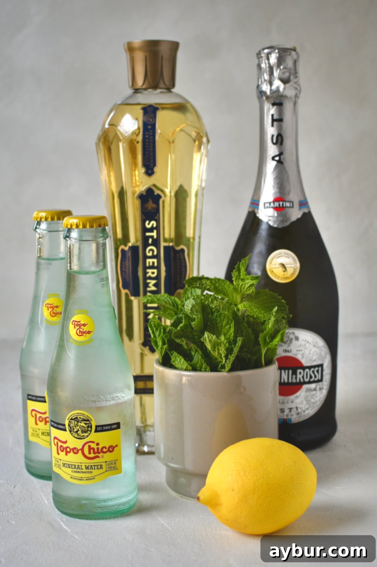 Ingredients needed to make Hugo Spritz, including elderflower liqueur, sparkling water, prosecco, fresh mint, and lemon.