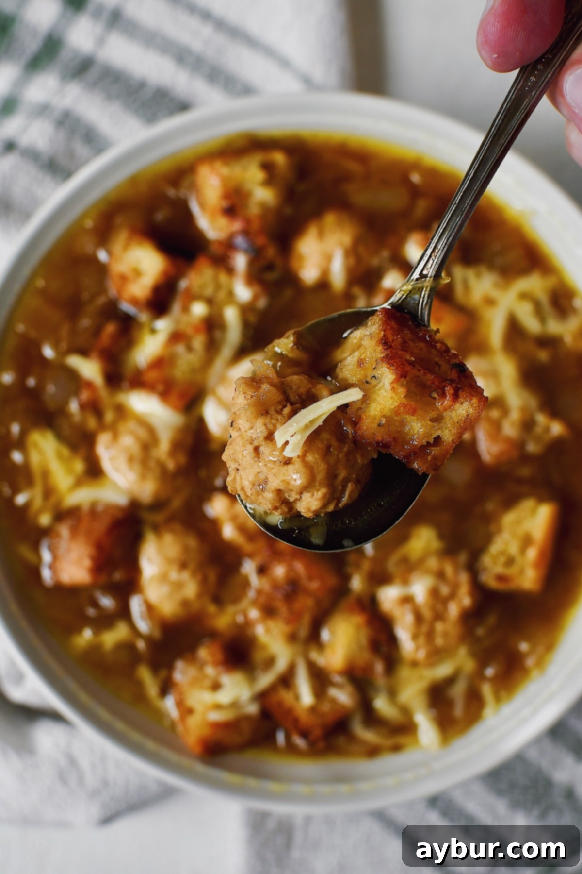 A spoon lifting a meatballs, crouton, and cheese from a bowl of Cheesy French Onion Soup, showcasing the rich texture and delicious additions.