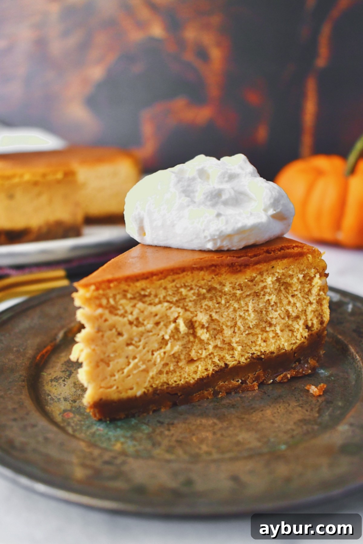 A single slice of pumpkin cheesecake sits in the foreground, with the beautiful full cheesecake in the background, both generously topped with whipped cream.